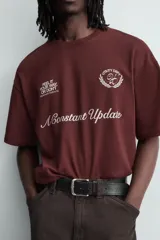 Camiseta de manga corta y cuello redondo, color granate oscuro, con estampado frontal en blanco que incluye texto y un escudo circular.