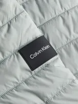 Campera acolchada de poliéster reciclado, color gris claro, con cuello alto y cierre frontal de cremallera negra. Presenta costuras horizontales y una insignia rectangular con el logo de Calvin Klein en el pecho.