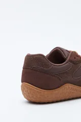 Zapatilla de estilo barefoot color marrón, confeccionada con una combinación de gamuza y materiales sintéticos. Presenta un diseño de perfil bajo con suela de goma flexible en color caramelo que incluye el logo de la marca grabado en el lateral.
