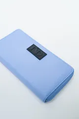 Billetera rectangular con cierre, color celeste pálido, con parche frontal negro que lleva las letras "CNJ" en relieve. Incluye un llavero de muñequera con cordón trenzado en rosa fuerte y verde lima.