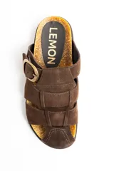 Sandalias tipo zueco de cuero gamuzado color marrón, con diseño de tiras cruzadas, puntera cerrada y una gran hebilla metálica dorada en el lateral. Cuenta con base de corcho y suela plana.