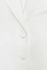 Blazer crop satinada de color blanco, con solapas, manga larga, cierre frontal con botones y detalle de abertura en la espalda.