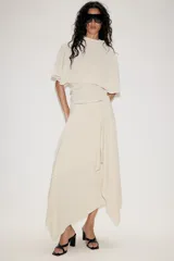 Top fluido de color beige con cuello perkins y mangas acampanadas por debajo del codo. Presenta un diseño de bajo redondeado y textura sutil en la tela.