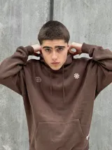 Canguro marrón con capucha ajustable con cordones, bolsillo tipo canguro y puños elásticos. Presenta un logo de un globo terráqueo y una flor bordados en el pecho.