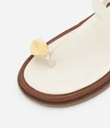 Sandalia plana tipo salomé color beige, con tiras finas, cierre con hebilla lateral y detalle decorativo dorado en la parte frontal. Cuenta con una suela de plataforma baja en tono madera.