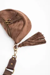 Cartera tipo bandolera de gamuza sintética color marrón, con diseño de media luna, cierre frontal con doble tirador metálico y detalle de borla lateral.
