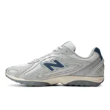 Championes New Balance modelo 204L, con diseño inspirado en el running de los años 2000. Presenta una capellada de ante y malla en tonos grises, con detalles en azul marino en el logo lateral y el talón. Cuenta con una suela de EVA con diseño técnico y líneas arqueadas.