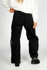 Pantalón de jean negro con corte recto, tiro alto, bolsillos delanteros y traseros, cierre con cremallera y botón. Presenta tres líneas blancas verticales a lo largo de la pierna y cierres metálicos dorados.