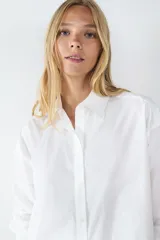 Camisa blanca de popelina de algodón, de corte amplio, manga larga, cuello clásico, cierre frontal con botones y puños abotonados.