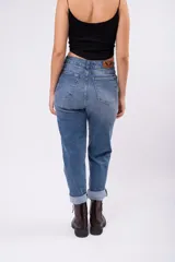 Pantalón tipo mom jean de tiro alto, confeccionado en denim rígido de color azul medio con efecto lavado. Presenta un corte recto y holgado con ruedos doblados, cierre frontal con botón y bolsillos laterales.