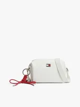 Cartera bandolera blanca Tommy Jeans de poliuretano, diseño cuadrado con cierre de cremallera y compartimento principal. Cuenta con correa ajustable, insignia metálica con el logo de la marca en el frente y un colgante lateral extraíble de cordón rojo trenzado con argollas metálicas.