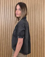 Camisa de denim de algodón celeste, con cierre frontal de botones, cuello alto con volados y mangas cortas abullonadas con puños elastizados fruncidos.