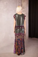 Falda larga de cuero verde oliva con diseño de paneles. La parte superior es de cuero liso con cierre frontal, decorada con un galón de borlas en tonos morados y verdes. La parte inferior presenta una falda de tela fluida con estampado floral y geométrico en tonos violeta, morado y verde.