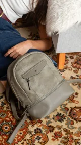 Mochila de diseño minimalista en color verde oliva, confeccionada en símil cuero. Presenta un bolsillo frontal con solapa y cierre, apertura superior con cierre y correas regulables para la espalda, además de un asa de mano superior.