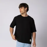 Remera oversize negra de lino y rayón entramado, con cuello redondo, manga corta caída y etiqueta decorativa en el borde inferior.