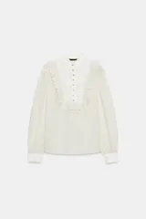Camisa de rayas finas color beige y blanco, con pechera frontal lisa en tono crudo, cuello mao y volantes aplicados alrededor de la botonadura y los hombros. Cuenta con mangas largas abullonadas y puños rectos a contraste.