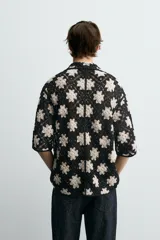 Camisa de manga corta con cuello solapa y cierre frontal de botones, confeccionada en tejido de crochet con patrón de flores en negro y beige.