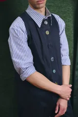 Mono largo de color negro con cierre frontal de botones vintage de distintos colores. Presenta un diseño sin mangas, con bolsillos laterales y una cinta para herramientas en la parte trasera.