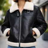Campera tipo aviadora United Uy de ecocuero negro, con forro interior, cuello, puños y ruedo de corderito blanco. Cuenta con cierre frontal de cremallera metálica, dos bolsillos ribeteados y detalles de hebillas en el cuello.