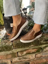 Zapatilla marrón de cuero con diseño entretejido, elástico lateral y suela blanca.