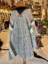 Poncho de lana virgen tejido en telar con diseño geométrico a rayas en tonos gris, marrón y celeste, con apliques de cuero en el escote y flecos en el ruedo.