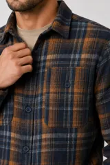 Sobrecamisa de franela a cuadros en tonos marrones, blancos y rojos, con mangas largas, bolsillos frontales con solapa y cierre de botones.