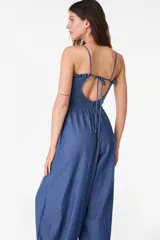 Mono largo de denim color celeste, sin mangas, con escote nido de abeja elástico y volado superior, tirantes finos para atar al cuello, perneras amplias de corte abombado con puños ajustables mediante lazos y bolsillos laterales.