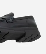 Zapato masculino tipo mocasín de color negro, confeccionado en poliuretano con efecto vinilo. Presenta punta redonda, detalle de antifaz y suela track gruesa con pespuntes en contraste.