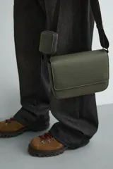 Bandolera verde militar con acabado engomado y textura geométrica. Tiene un compartimento principal con cierre de solapa y botón a presión, un bolsillo interior con cremallera y un asa de hombro ajustable con monedero.