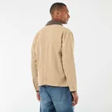 Chaqueta de trabajo (chore coat) color beige, confeccionada en tejido de algodón tipo sarga o drill, con cuello contrastante de pana marrón oscuro. Presenta cierre frontal de botones metálicos y cuatro bolsillos frontales de parche (dos superiores y dos inferiores).