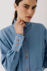 Camisa de jean azul con cuello mao con volados, cierre frontal con botones de madera y mangas largas con puños con volados.