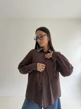 Camisa oversize de color marrón con cuello camisero y puños con detalle de bordado festoneado en color blanco. Presenta cierre frontal de botones y mangas largas.