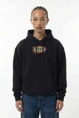 Hoodie negro de corte relajado con capucha y cordones ajustables. Presenta un estampado gráfico central en el pecho con forma ovalada y bordes irregulares en tonos verde y rojo.