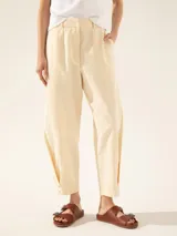 Pantalón color crema de tiro alto, tipo bombe, con recortes verticales en el delantero, pliegues en la bota con broche níquel, cintura con gancho pantalonero y botón interno, bolsillos ojal y pinza de entalle en la espalda.