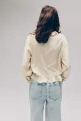 Blusa de viscosa color amarillo claro con detalles de broderie, cuello con volados y abertura frontal en V con tirantes finos. Presenta mangas globo largas y ribetes con volados en los puños y el dobladillo.