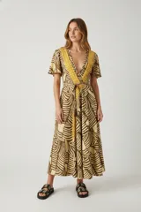 Vestido midi cruzado con mangas cortas, estampado de hojas en tonos marrón y amarillo mostaza. Presenta un profundo escote en V con borde decorativo bordado en amarillo mostaza y lazo ajustable a la cintura.