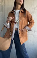 Chaqueta de gamuza color camel, con corte recto, cuello camisero, cierre frontal con botones metálicos y bolsillos frontales con solapa.