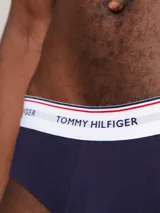 Pack de tres calzoncillos tipo slip de la marca Tommy Hilfiger, confeccionados en algodón elástico, con cintura elástica con el nombre de la marca y líneas distintivas. Incluye una unidad con cintura blanca, otra con cintura azul marino y otra con cintura roja.