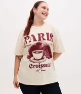 Remera oversize de algodón color beige, de cuello redondo y manga corta, con gran estampado frontal en tonos bordó con temática de París, incluyendo las inscripciones 'PARIS', 'France', 'Croissant & Crème' y un diseño gráfico de una taza de café y un croissant.