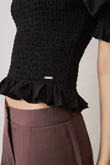 Blusa negra con cuerpo fruncido tipo smock, mangas cortas abullonadas con puños elásticos y volados en el cuello y el ruedo.