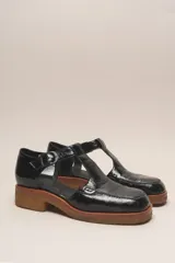 Zapato tipo guillermina de cuero charol color marrón oscuro, con tira T y hebilla ajustable en el lateral. Posee puntera cuadrada y suela de goma color caramelo con taco bajo.