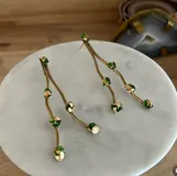 Aretes largos colgantes hechos a mano en bronce, con forma de rama o tallo ondulado. Presentan apliques de piedra verde esmaltada con detalles de pan de oro o lámina dorada incrustada.