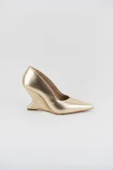 Zapato de cuero metalizado en color dorado, con punta fina y taco cuña escultural de diseño geométrico.