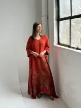 Vestido largo color terracota con escote en V, mangas tres cuartos y diseño amplio, decorado con un estampado floral grande en tono marrón en la parte inferior.