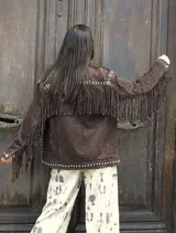 Chaqueta de gamuza color chocolate, con diseño de flecos en el frente y la espalda, decorada con tachas metálicas en cuello, hombros y puños. Cuenta con cierre frontal de botones y forro interior.