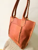 Cartera tipo shopper de cuero genuino marrón, con terminaciones artesanales trenzadas en las asas y los laterales, bolsillo frontal externo y asas largas para llevar al hombro.