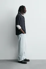 Camisa de algodón negro, de corte relajado (relax fit), con cuello bowling, manga corta y bolsillo plastrón en el pecho. Cierre frontal con botones a presión.