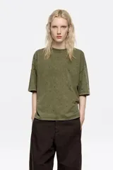 Camiseta de corte boxy en color verde oliva con efecto lavado, cuello redondo y manga corta. Presenta pequeños remaches metálicos distribuidos por toda la prenda.