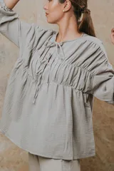 Camisa de algodón con textura arrugada, cuello redondo con cierre de cordones y mangas largas con puños elásticos. Presenta un diseño holgado con frunces en el pecho.
