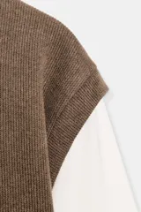 Prenda de manga larga blanca con un chaleco de tejido de punto azul marino superpuesto, creando un efecto de dos piezas. El chaleco es de cuello redondo y corte recto.